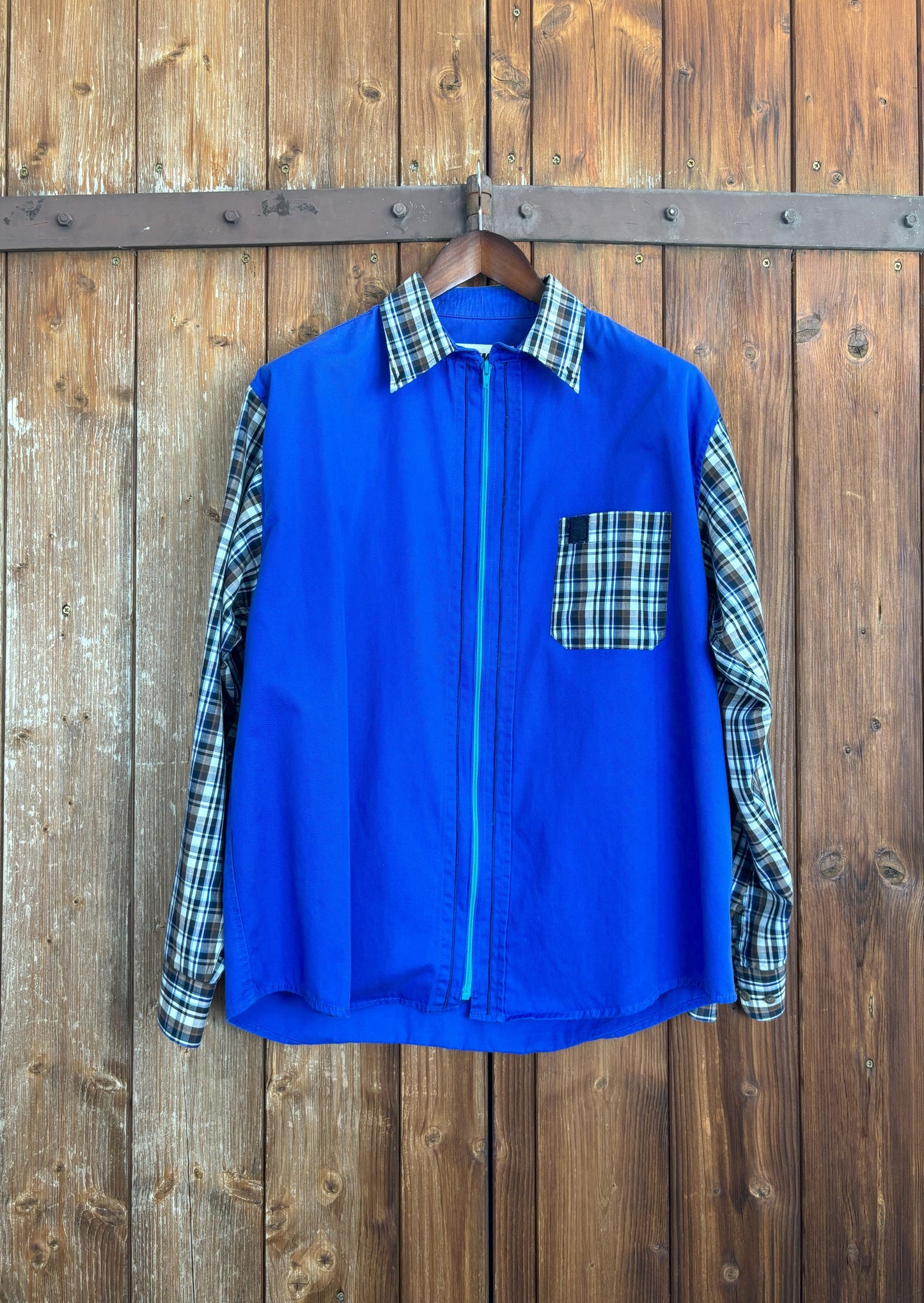 Qualitativ hochwertiges, blaues Hemd mit türkisfarbenem Reißverschluss anstelle der Knopfleiste. Kragen, Ärmel und Brusttasche wurden durch Teile eines blau-braun-weiß karierten Hemdes ersetzt. Mit Kentkragen. 